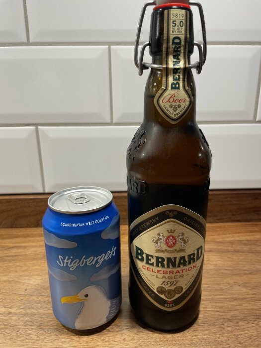 En ölburk från Stigbergets och en glasflaska med Bernard Celebration Lager står på en träbänk framför vit kakelvägg.