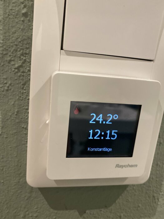 Termostat av märket Raychem med en digital display som visar 24,2 grader och klockan 12:15, installerad på en vägg med grön färgtextur.