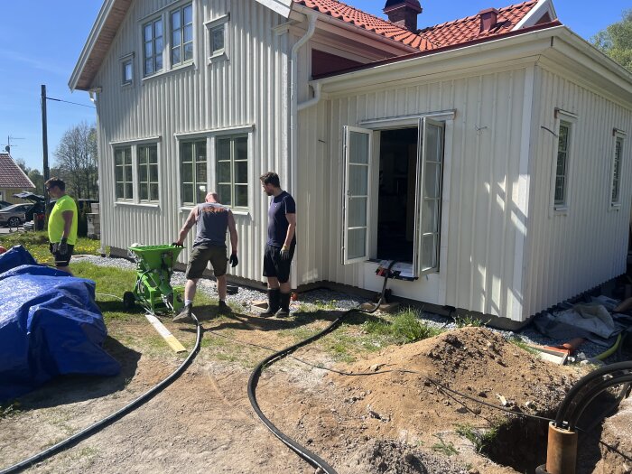 Tre personer arbetar på en byggarbetsplats utanför ett ljusgult hus. Jordhögar och rör syns på marken, och en öppen dörr leder in i huset.