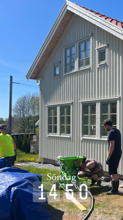 Tre personer arbetar på en byggarbetsplats utanför ett vitt trähus med gröna fönster. En person lutar sig över en grön maskin. Texten "Söndag 14:50" syns.