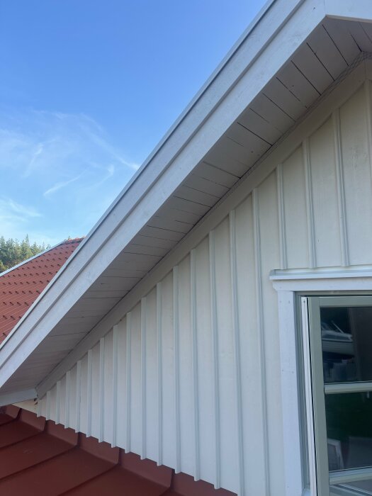 Ett närbildsfoto av husets yttervägg med vit träpanel och ett fönster, samt takfot och en del av det rödbruna taket, mot en klarblå himmel.