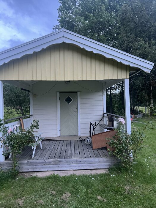 Framsida av ett ljust träfärgat hus med en veranda och dekorativ gul panel i överkant, omgiven av träd och gräsmatta.