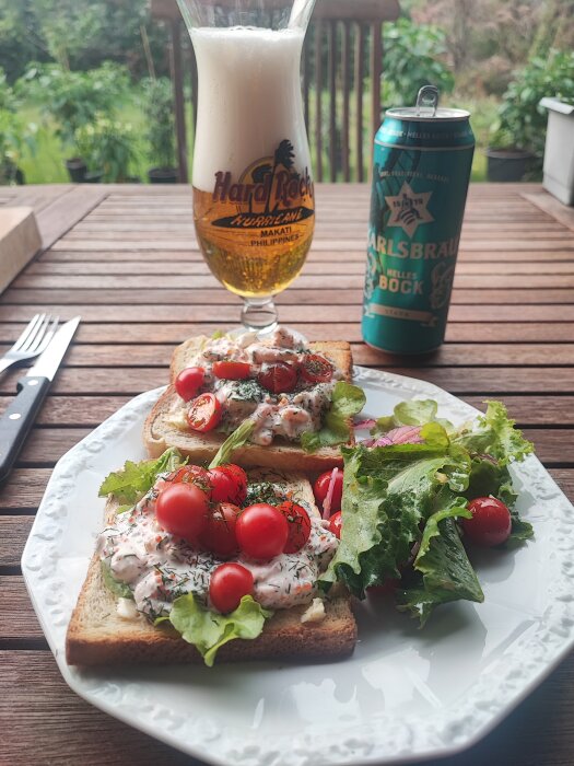 Två brödskivor med Skagenröra och körsbärstomater på en tallrik, bredvid en sallad. I bakgrunden ett glas öl och en ölburk på ett trädäck.