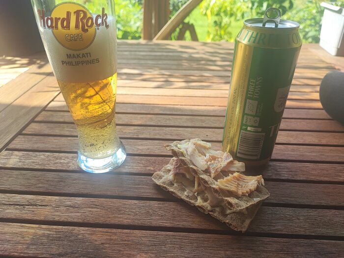 Ett glas med öl från Hard Rock Cafe och en burk öl på ett träbord tillsammans med rökt abborre på knäckebröd i solskenet.