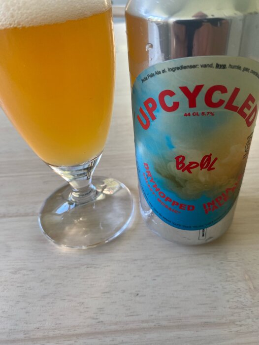 Konservburk med öl märkt "Upcycled Bröl" bredvid ett glas fyllt med gyllene öl med ett vitt skumtäcke på ett träbord.