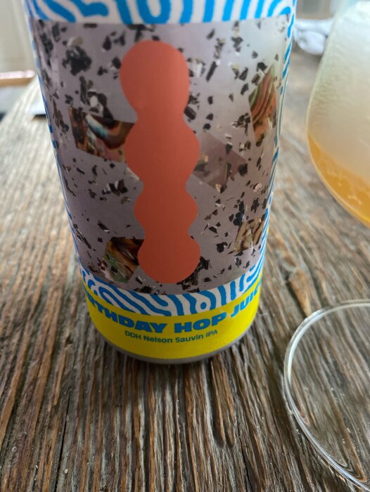 Ölflaska med etiketten "Birthday hop juice DDH Nelson Sauvin IPA" och orange grafisk design, samt glas med öl på ett träbord.