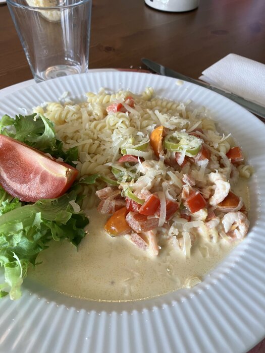 Tallrik med pasta och räksås toppad med parmesan, tillsammans med en sallad av gröna blad och en tomathalva. Ett glas vatten i bakgrunden.