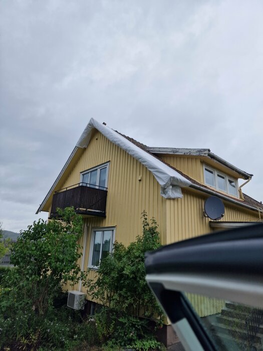 Gul villa med ett mörkt balkongräcke och plastskydd över taket, troligen under renovering, med buskar framför och en parabolantenn på väggen.