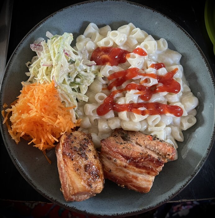 En tallrik med stuvade makaroner toppade med ketchup, grillat sidfläsk, rårivna morötter och coleslaw.