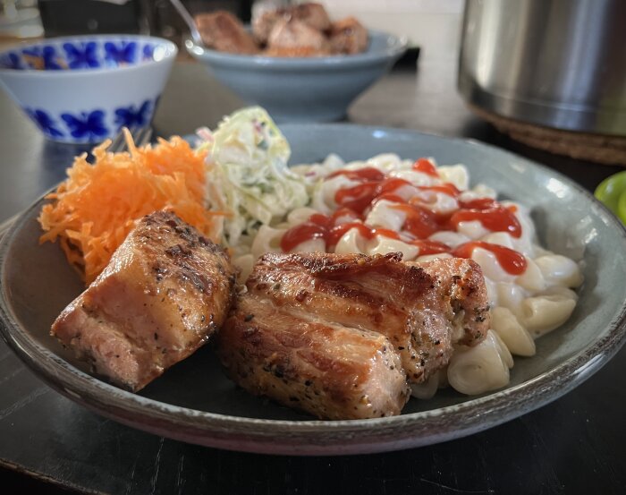 En tallrik med stuvade makaroner och ketchup, grillat sidfläsk, rårivna morötter och coleslaw. I bakgrunden syns en blåvit skål och mer grillat sidfläsk.