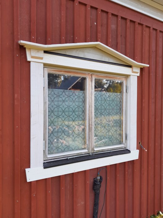 Fönster med ny monterad vit karm på en rödfärgad fasad, spetsgardiner täcker glaset och en kabel hänger bredvid.