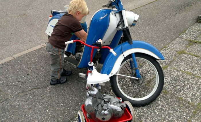 Ett litet barn i brun tröja och grå byxor står intill och arbetar med en blå Puch Montana-moped. En liten röd leksaksmotorcykel står bredvid dem.