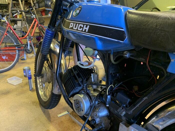 Blå Puch Montana-moped under reparation i ett garage, med två cyklar och verktyg synliga i bakgrunden.