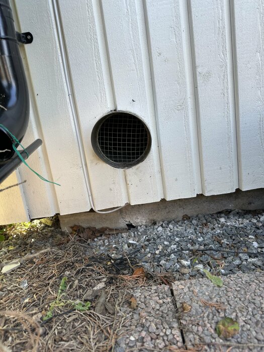 Cirkulärt ventilationsutblås med galler på en vit husfasad över en grus- och markyta, inklusive en del av en svart rörinstallation.