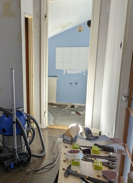 Renovering av liten toalett med verktyg och dammsugare i förgrunden, och en blåmålad vägg med bortplockat kakel och trasigt golv i bakgrunden.