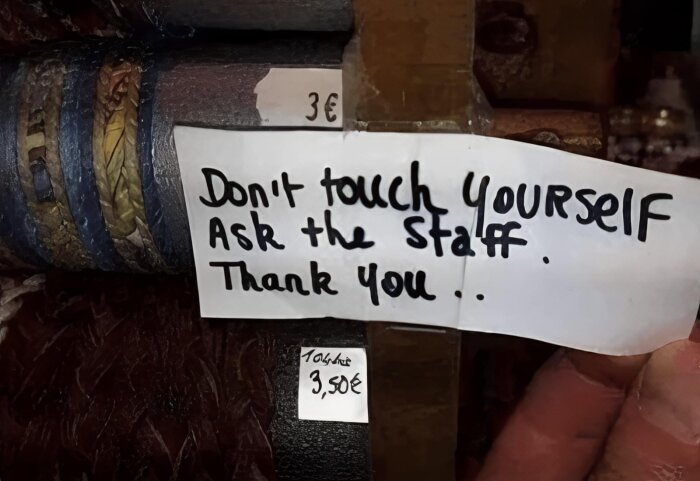 Lapp med texten "Don't touch YOURSELF Ask the Staff Thank you" är fastsatt på en träribba med prislappar på 3€ och 3,50€ i en butik.