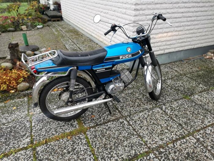 Blåmetallic Puch Montana-moped parkerad på en stenlagd uteplats bredvid en vit vägg och omgiven av höstväxter.