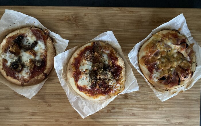 Tre små pizzor med olika toppings på en träskärbräda, med mozzarellaost och ingredienser som salami, skinka, tomat, kapris, parmesan och cheddar-texmexost.