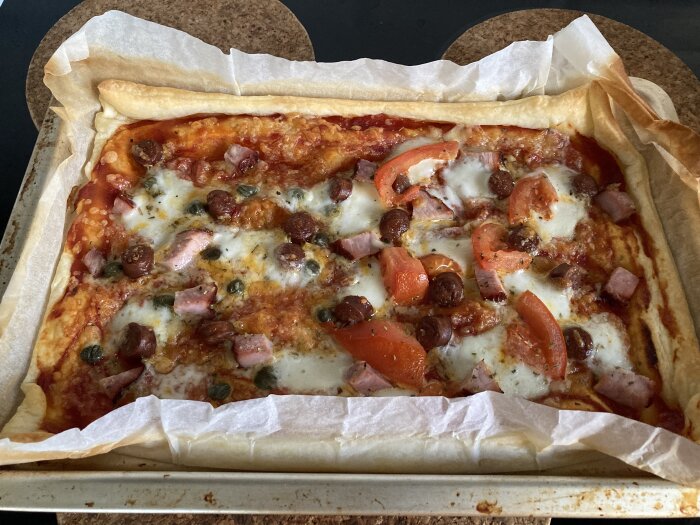 Fyrkantig pizza på bakplåtspapper med topping som tomater, salami, skinka, kapris, mozzarella och tomatsås, bakad i ugn.