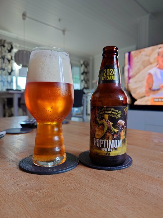 Ett glas fyllt med en orangefärgad öl med skumkrona bredvid en flaska Hoptimum Triple IPA från Sierra Nevada på ett bord med underlägg.