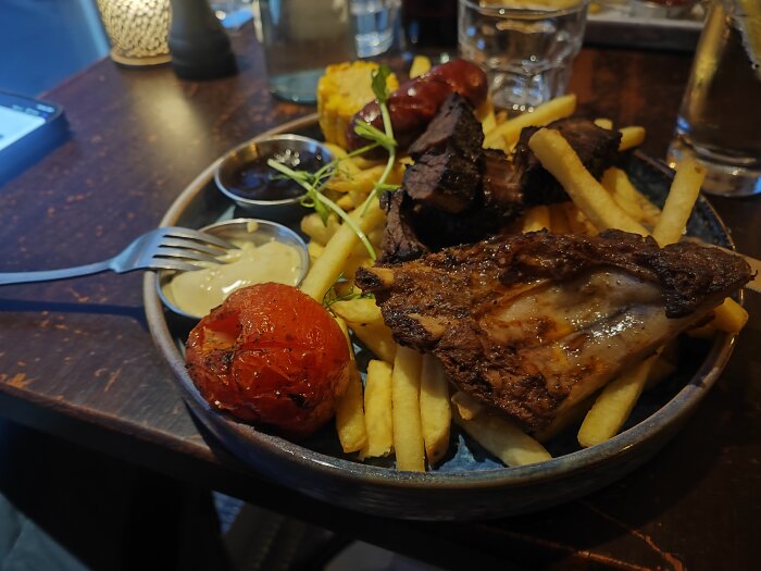 Tallrik med mixed grill på en restaurang, innehållande pommes frites, revbensspjäll, grillad korv, såser och en grillad tomat.