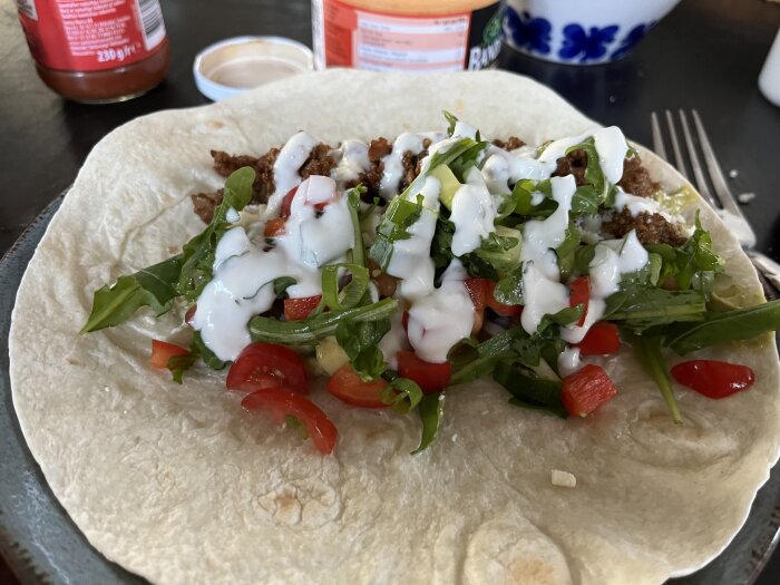 En tortilla fylld med nötfärs, sallad, tomater och vitlökssås med burkar av salsa och ostsås i bakgrunden.