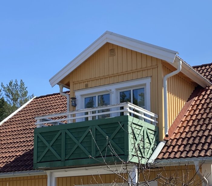 Gult hus med tegeltak och grön balkong, där balkongen har vita räcken och är täckt med gröna plankor, fotograferat under en klarblå himmel.