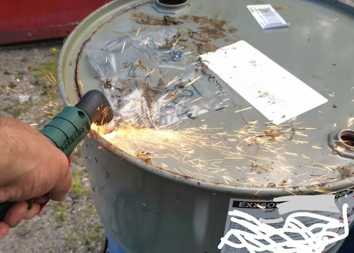 Hand använder en grön slipmaskin för att bearbeta en metalltunna, vilket skapar gnistor runt verktyget.