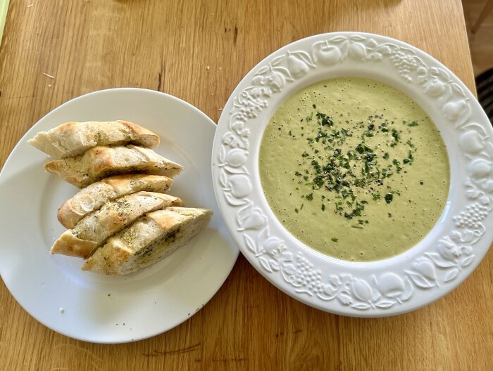 En tallrik med potatis- och broccolisoppa toppad med örter bredvid en tallrik med skivor av vitlöksbröd.