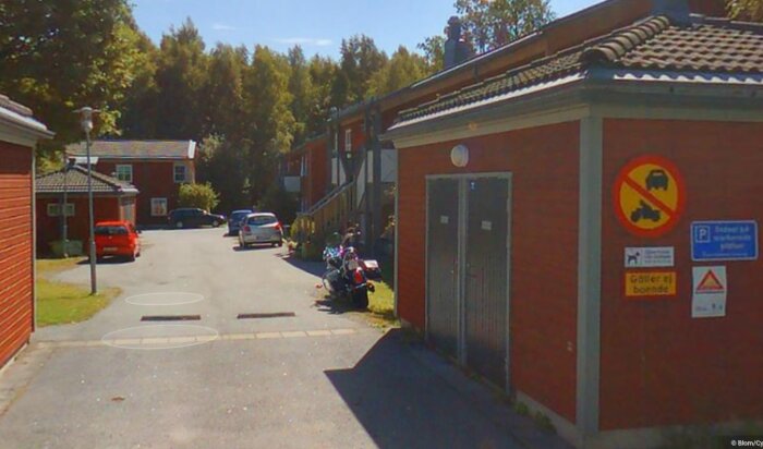 Bilden visar en gata i ett radhusområde med parkerade bilar och en motorcykel. Skyltar på en byggnad varnar för lekande barn och visar parkeringsrestriktioner.