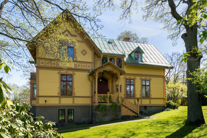 Gul villa med gröna tak och bruna detaljer, omgiven av frodig grönska och träd. Huset påminner färgmässigt om Villa Villekulla.