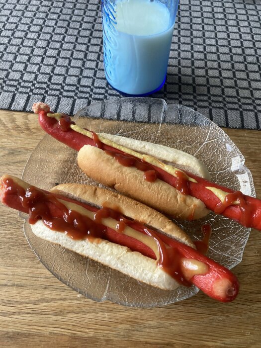 Två varmkorvar med senap och ketchup på ett glasfat, bredvid ett glas mjölk på en bordsduk.