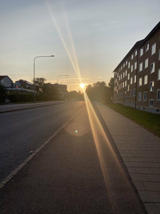 Solnedgång över en tom gata, med gatubelysning och bostadshus på ena sidan. Solens strålar lyser längs trottoaren och bildar ett vackert ljusspel.