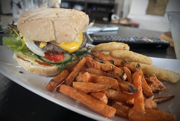 Tallrik med hamburgare, sötpotatispommes och lökringar. Hamburgaren har grönsaker, picklad lök, guacamole och ost. Coleslaw ingår också men syns ej på bilden.