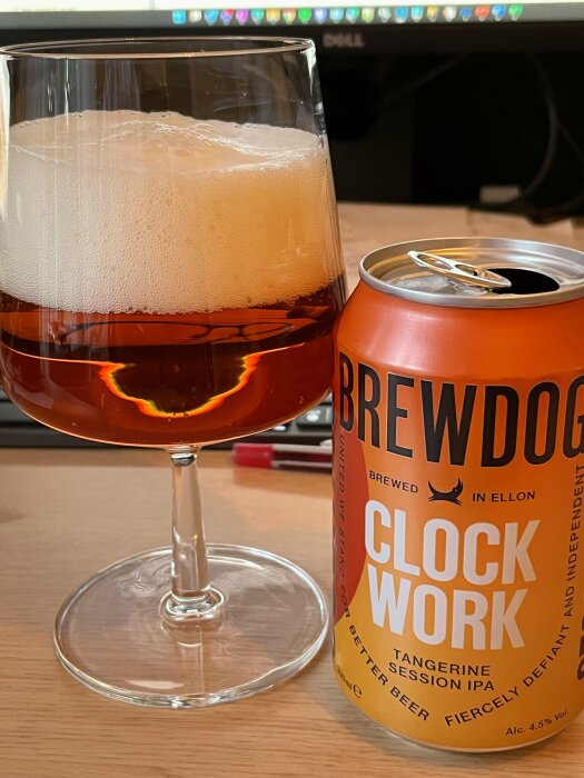 Ett glas öl med skum på toppen bredvid en burk Brewdog Clockwork Tangerine Session IPA på ett skrivbord.