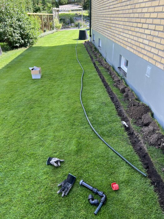 Gräsmatta med nylagd rörledning för automatisk bevattning, grävningsarbete längs husväggen, samt handskar och kopplingar på gräset.