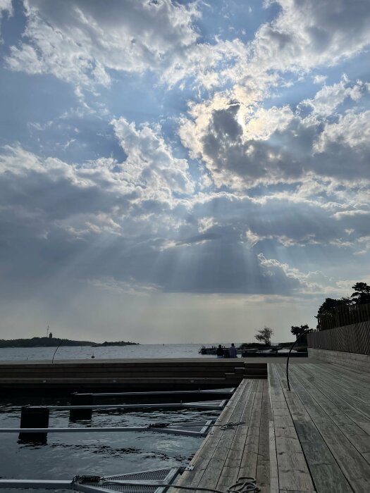 En brygga vid havet med solstrålar som skiner genom molntäcket på himlen. Två personer sitter på bryggan och tittar ut över vattnet.