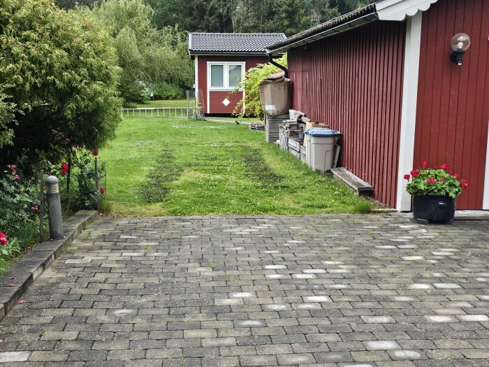 Grön gräsmatta med en öppning på 3,6 meter mellan ett rött garage och en häck. Tegelbelagd uppfart och en blomkruka med röda blommor i förgrunden.