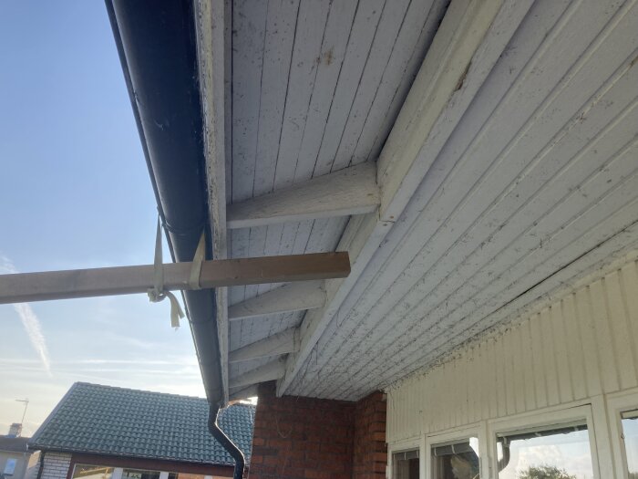 Pergola fäst vid en hängränna under ett tak utanför ett hus med en vitmålad träfasad och tegelvägg.