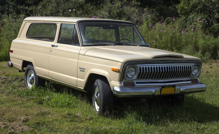 En beige 1970-tals Jeep Cherokee Chief parkerad på gräs med blommor och grönska i bakgrunden.