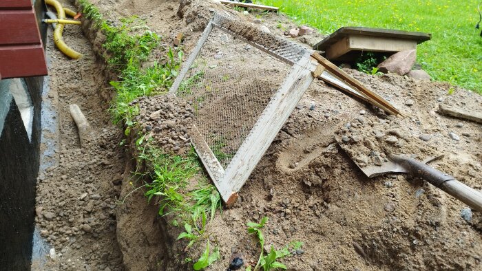 Utgrävd mark med grävt dike vid husgrund, ett nät placerat över dikeskanten och några verktyg som spade och slangar på marken.