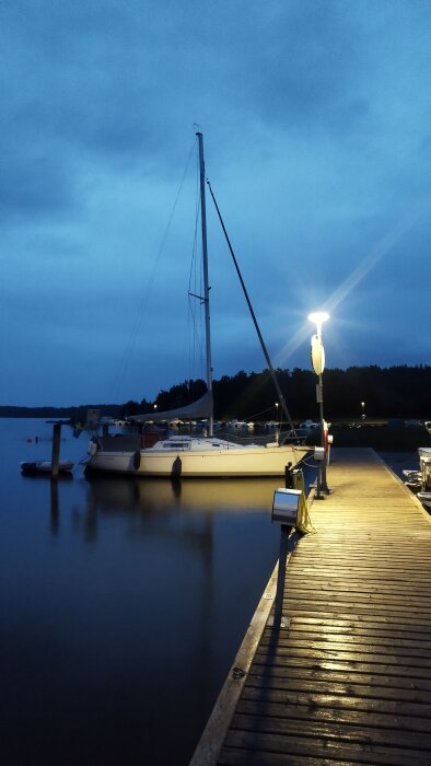 En segelbåt förtöjd vid en upplyst brygga under kvällstid med molnig himmel i bakgrunden och mörka silhuetter av träd i fjärran.