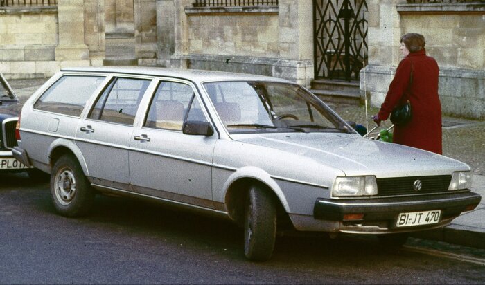 En Volkswagen Passat parkerad vid trottoaren bredvid en äldre kvinna i röd kappa med en grön påse i en europeisk stad.