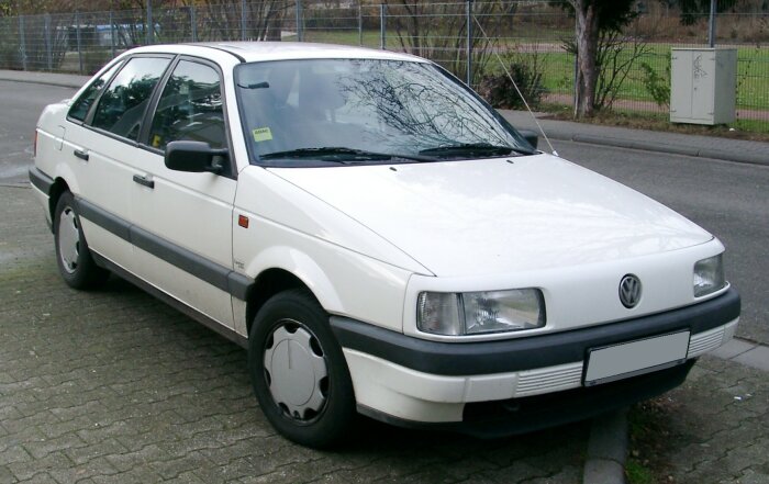 En vit Volkswagen Passat från 1980-talet parkerad vid en trottoarkant med ett staket och grönt område i bakgrunden.