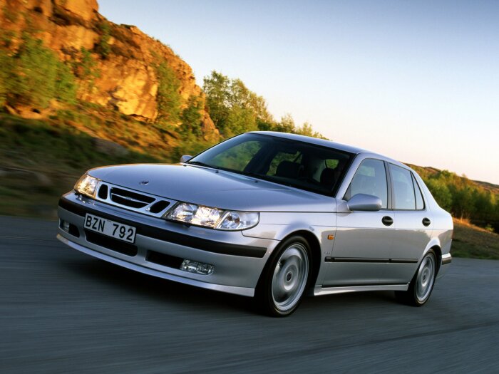 En silverfärgad Saab 9-5 bil från 1998 kör längs en väg med natur och klippor i bakgrunden.