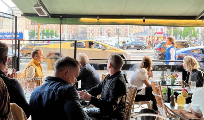 Flera personer sitter på en uteservering vid Strandvägen i Stockholm medan en Tesla Cybertruck kör förbi utanför.