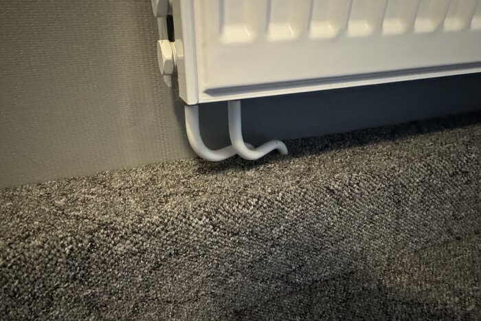 Kurvig böj på radiatorrör som är installerade nära en vägg och ovanför en grå matta.