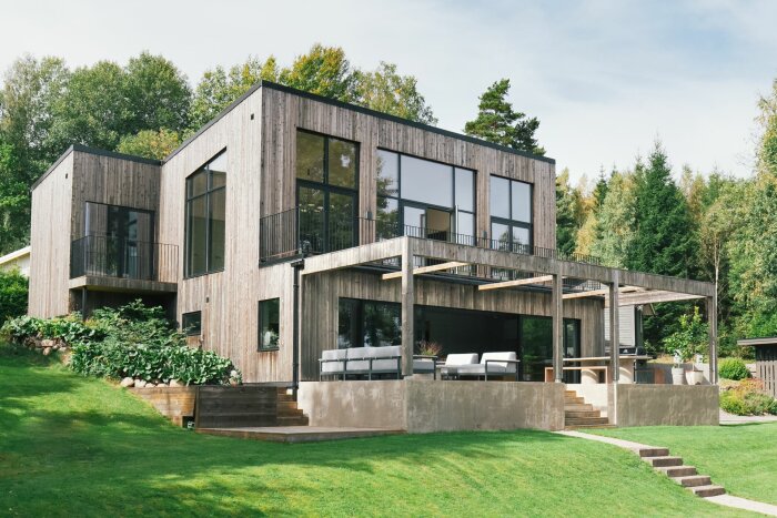 Modernt hus i två våningar med stora fönster och träfasad, omgiven av grönska och uppvuxna träd. Stenlagd uteplats med sittplatser under en pergola.