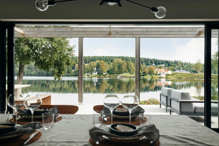 Utsikt från matplats med dukat bord över en sjö, med träd och hus vid strandkanten och skog i bakgrunden.