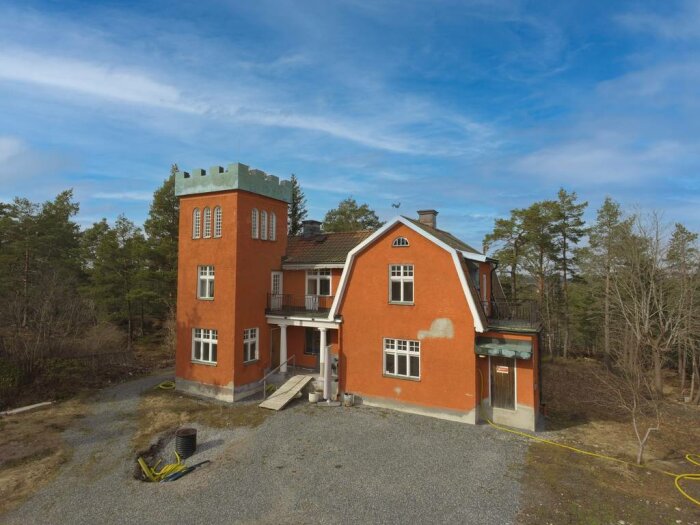 Stort orangefärgat hus med ett torn till vänster, beläget i en skogsmiljö under en blå himmel.